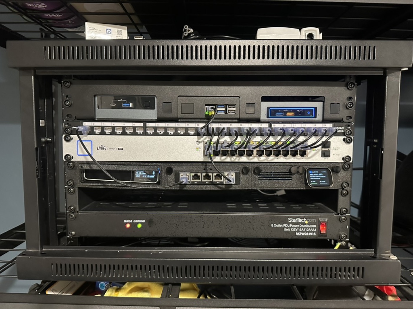 A compact home network rack with a UniFi switch, patch panel, mini PCs, and a rack-mounted power strip.