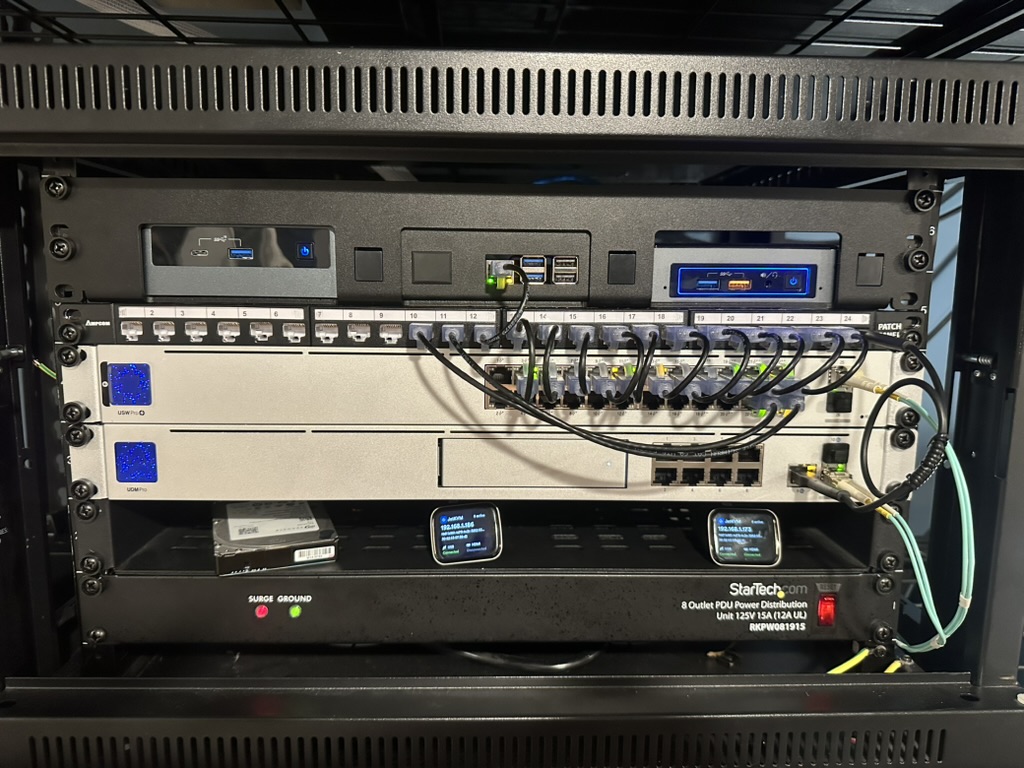 The current home rack: two Intel NUCs and a Raspberry Pi, a patch panel, a UniFi Switch Pro 24 PoE and a UniFi Dream Machine Pro, above a shelf with an external hard drive, two JetKVMs and a StarTech power strip.